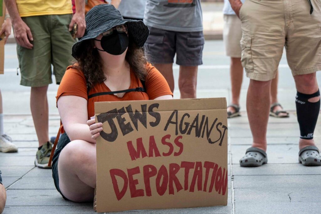 Demonstrator wearing a hat and face mask holds a cardboard sign reading, "Jews Against Mass Deportation". 